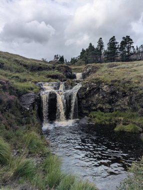 Yosun üzerine çağlayan bir şelale Glen Brittle 'daki Peri havuzunda kayaları yansıtıcı bir havuza dönüştürdü. Çimenli yamaçlar ve kozalaklı ağaçlar bulutlu bir gökyüzü altında Bu uzak İskoçya manzarası