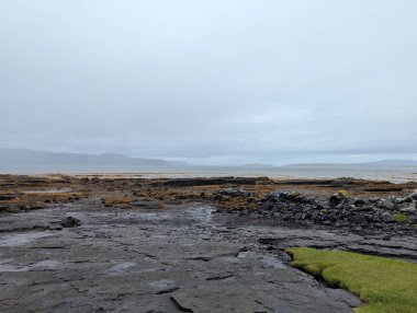 Katmanlı kaya platformları ve gelgit havuzları, Skye Adası 'ndaki Broadford kıyı şeridi boyunca uzanan çimenli arazilerle kesişiyor. Sisli bulutlu koşullar ve dağınık kıyı kulübeleri bu engebeli Highland deniz manzarasını oluşturur..