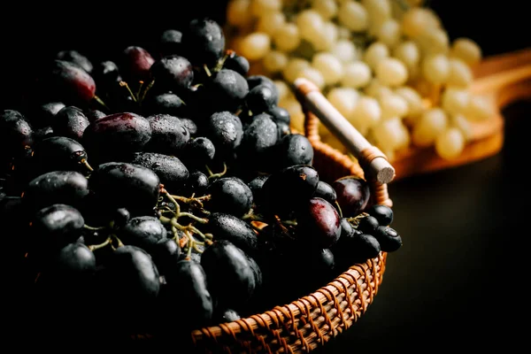 Arka planda taze yeşil üzümlerle dokuma sepetinde olgun siyah üzümler.