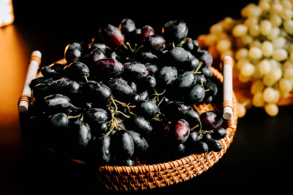 Arka planda yeşil üzümlerle dokuma sepetindeki olgun siyah üzümler.