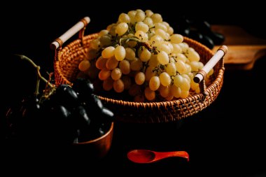 Siyah üzümlerin yanında sepetlenmiş olgun üzümler ve karanlık bir masada tahta bir kaşık.