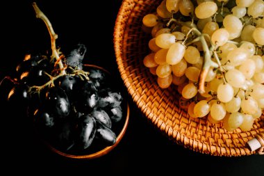 Olgun siyah ve beyaz üzümler, koyu bir yüzeyde dokuma sepetinde dizilmiş.