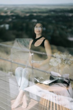 Güneşli bir günde manzaraya bakan pencerenin yanında düşünceli bir şekilde oturan kadın.