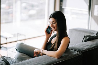 Modern bir oturma odasında rahatça otururken telefonda konuşan bir kadın.