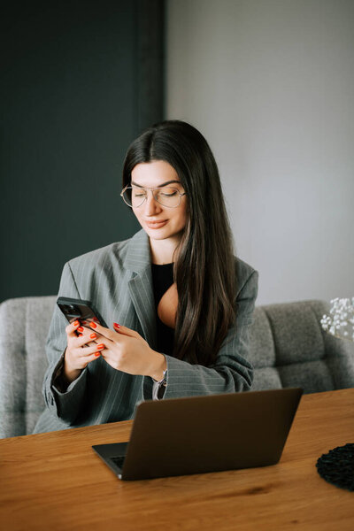 Женщина в очках проверяет смартфон, сидя за столом с ноутбуком