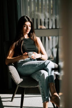 Kadın, güneşli odada modern sandalyede dinlenirken kahveden zevk alıyor.