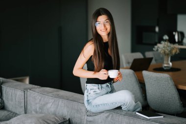 Modern bir yaşam alanında bir kanepede oturmuş bir fincan kahvenin tadını çıkaran bir kadın.