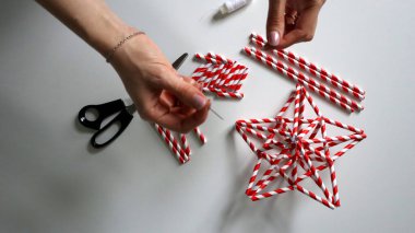 El yapımı. Kadın eli iplikle iğneyi tutuyor. Kokteyller için kağıt tüplerden Noel yıldızları yaratma sürecinde. Kırmızı, beyaz çizgili tüpler. Makas, beyaz iplik. Beyaz arka plan. Fotoğraf.