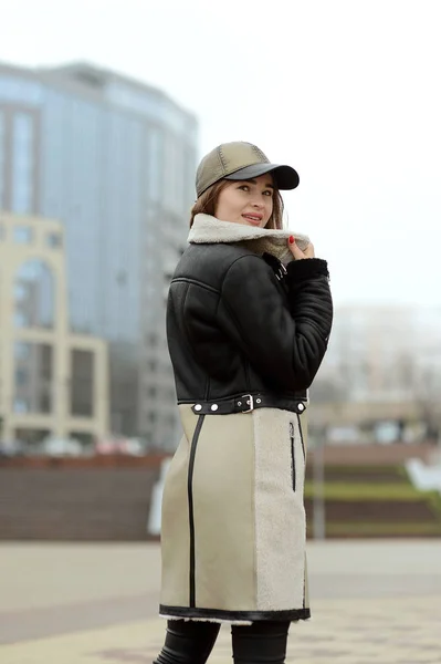 A girl in a black leather jacket and beige skirt in a cap posing in the city square — Stock Photo, Image