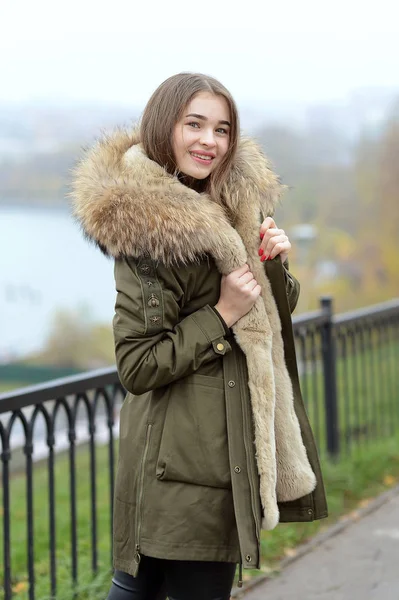 A girl in a green jacket with a fur collar in glasses posing near the lake — Stock Photo, Image