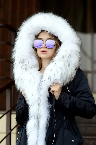 Una ragazza in giacca nera con un collo di pelliccia bianca e occhiali in posa per strada Foto Stock