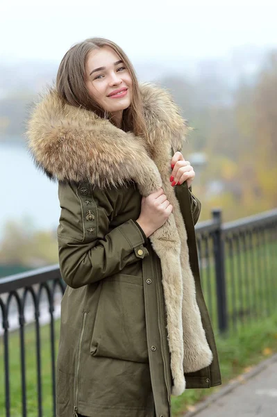 A girl in a green jacket with a fur collar in glasses posing near the lake — Stock Photo, Image