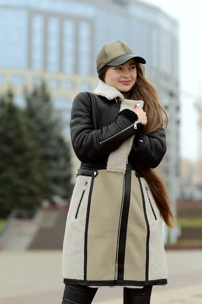 A girl in a black leather jacket and beige skirt in a cap posing in the city square — Stock Photo, Image