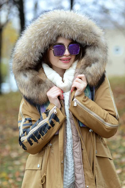 girl in a down jacket with fur and blue glasses walking in the woods