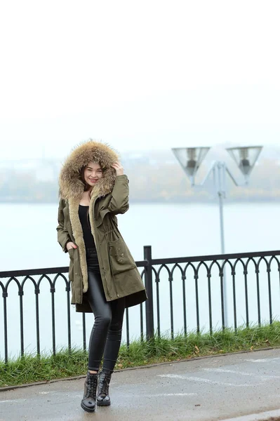 A girl in a green jacket with a fur collar in glasses posing near the lake — Stock Photo, Image