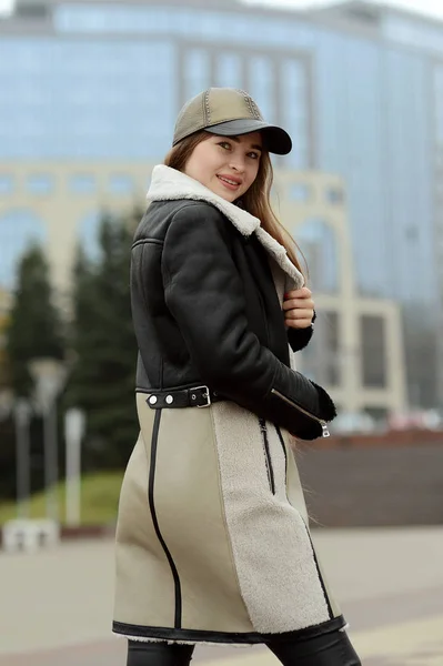 A girl in a black leather jacket and beige skirt in a cap posing in the city square — Stock Photo, Image