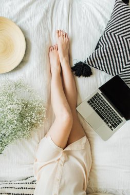 Yatakta dizüstü bilgisayarda çalışan genç kadın. Yaşam tarzı kompozisyon yukarıdan manzaralı dekore edilmiş beyaz çiçekler Buket, yastık ve ahşap plaka ile.