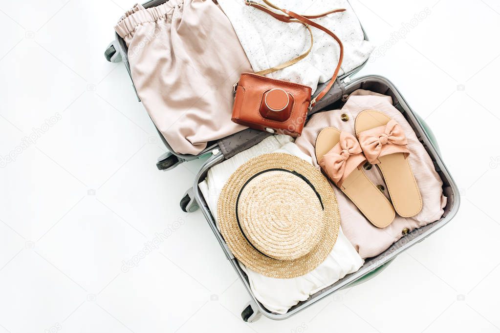 Hand luggage with stylish femininine clothes and retro camera on white background. Flat lay, top view. Summer fashion clothes concept.