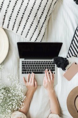 Yatakta dizüstü bilgisayarda çalışan genç kadın. Yaşam tarzı kompozisyon yukarıdan manzaralı beyaz çiçekler buket, yastık, şapka, cep telefonu ve ahşap plaka ile dekore edilmiştir. Serbest meslek ya da moda blogger ev çalışma alanı kavramı.
