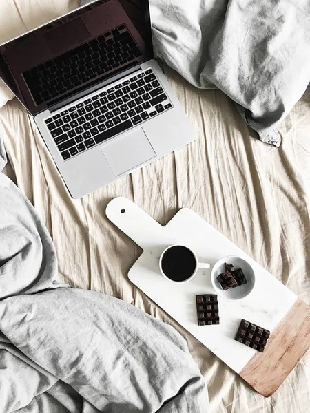 Laptop, pastel linens, breakfast with coffee and chocolate on marble cutting board. Flat lay, top view lifestyle concept.