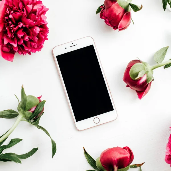 Smartphone in frame of pink peonies flowers on white background. Flat lay, top view mock up.