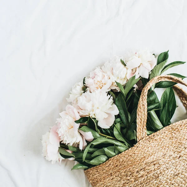 Straw bag with white peony flowers on white background. Flat lay, top view summer floral concept.