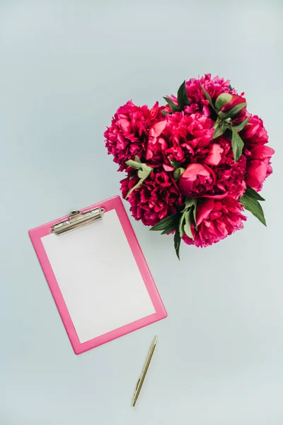 Minimalist flat lay office table desk with clipboard, peony flowers bouquet on blue background. Top view work concept mock up.