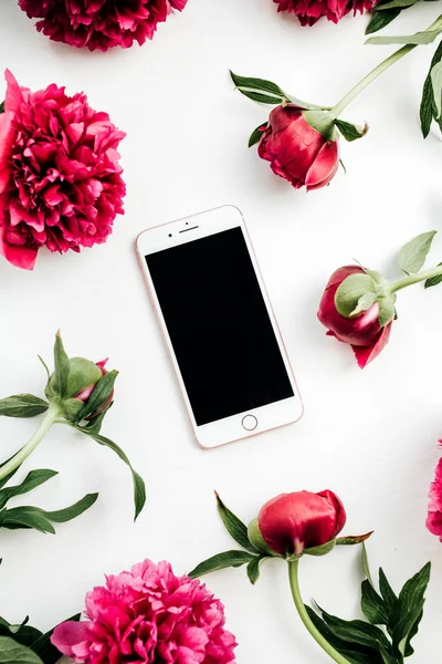 Smartphone in frame of pink peonies flowers on white background. Flat lay, top view mock up.