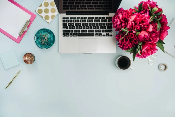 Workspace with laptop and pink peony flowers bouquet on blue background. Flat lay, top view home office desk.