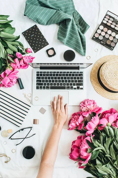 Flat lay, top view feminine office desk workspace with laptop, peony flowers, glasses, straw hat, cosmetics, accessories. Woman working on computer. Work concept.