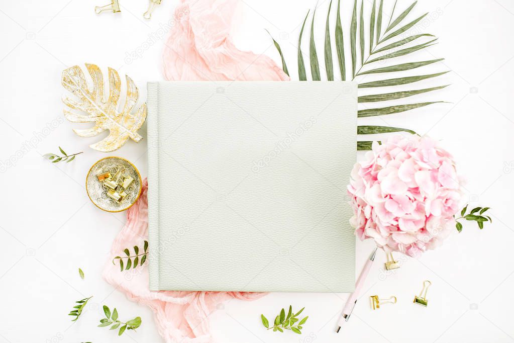 Composition with wedding or family photo album, hydrangea flower bouquet, tropical palm leaf, pastel pink blanket, gold monstera plate on white background. Flat lay, top view still life concept.