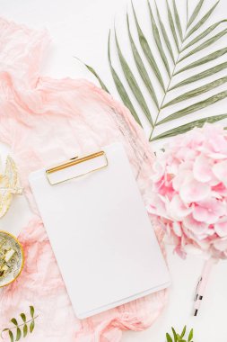 Modern home office desk workspace with blank paper clipboard, pink hydrangea flowers bouquet, tropical palm leaf, pastel blanket, monstera leaf plate and accessories on white background. Flat lay, top view rose gold mockup.