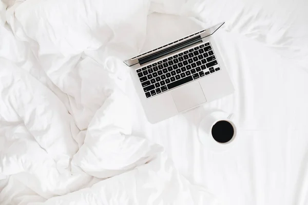 Flat lay, top view workspace in bed. Laptop and coffee cup in bed on clean white linens. Feminine work concept.