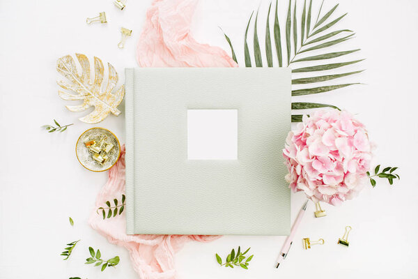Composition with wedding or family photo album, hydrangea flower bouquet, tropical palm leaf, pastel pink blanket, gold monstera plate on white background. Flat lay, top view mockup concept.
