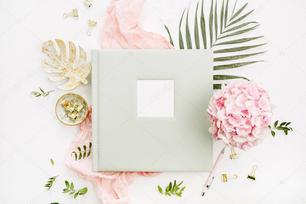 Composition with wedding or family photo album, hydrangea flower bouquet, tropical palm leaf, pastel pink blanket, gold monstera plate on white background. Flat lay, top view mockup concept.