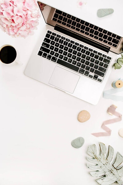 Modern home office desk workspace with laptop, pink hydrangea flowers bouquet, coffee cup, monstera leaf plate and accessories on white background. Flat lay, top view.