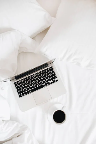 Laptop and coffee in bed with white linens. Freelance blogger workspace flat lay. Top view minimal background.