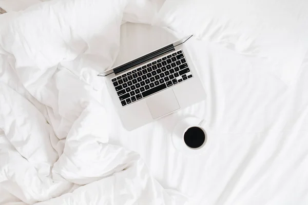 Flat lay, top view workspace in bed. Laptop and coffee cup in bed on clean white linens. Work at home concept.