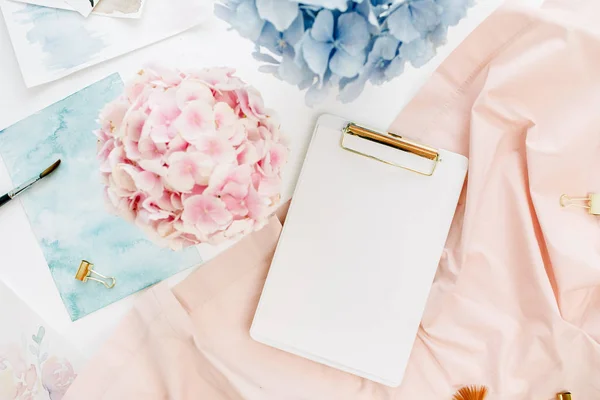 Art composition home office desk workspace with copy space clipboard, pastel hydrangea flowers bouquet, watercolor painting, peachy blanket. Flat lay, top view.