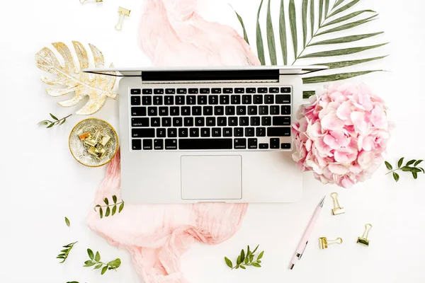 Woman home office desk with laptop, pink hydrangea flowers bouquet, tropical palm leaf, pastel blanket, monstera leaf plate and accessories on white background. Flat lay, top view rose gold workspace.