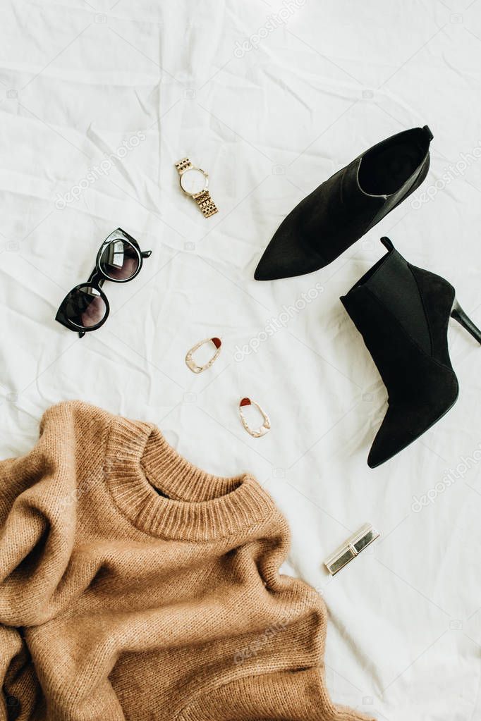 Flat lay, top view fashion concept. Trendy woman clothes and accessories on white blanket. Woolen sweater, boots, sunglasses, watch, earrings, lipstick.