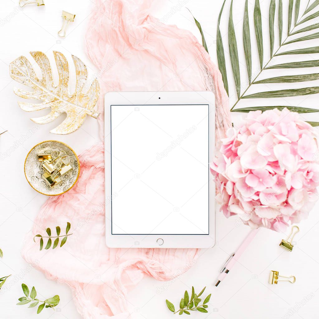 Blank screen tablet, pink hydrangea flowers bouquet, tropical palm leaf, pastel blanket, monstera leaf plate and accessories on white background. Flat lay, top view rose gold home office desk workspace mockup.