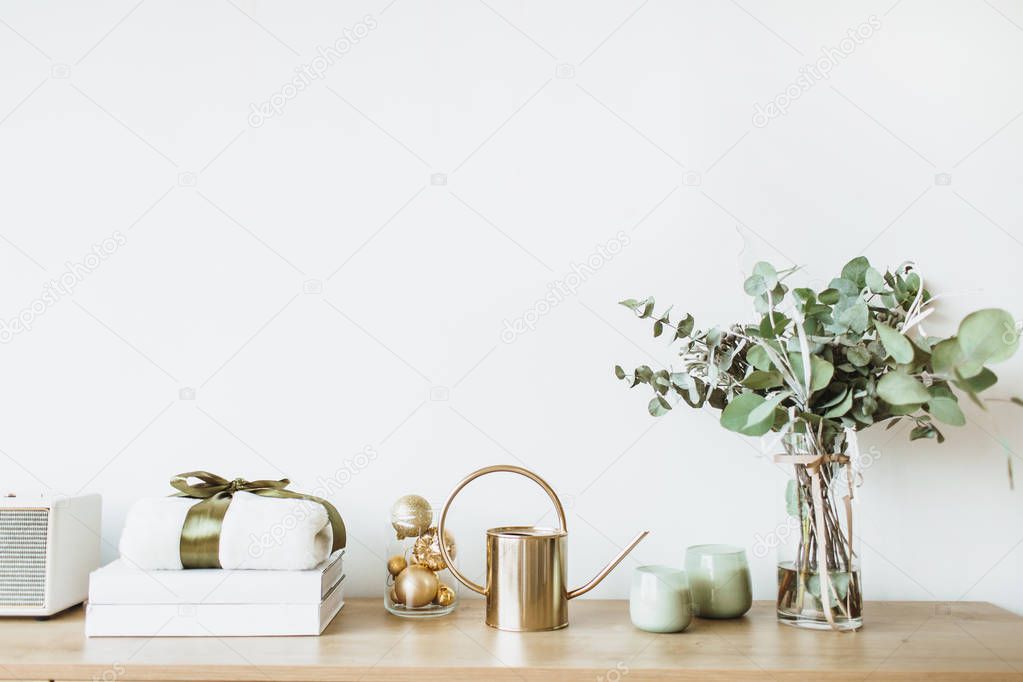 Modern interior design concept. Minimal Nordic Scandinavian style living room with wooden table with gift box, floral bouquet with eucalyptus, candles, radio at white wall. Pastel apartment for rent.