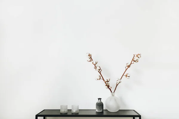 Modern scandinavian interior design concept. Stylish rack with cotton branch in vase, candles at white background.