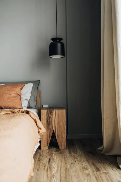 Modern Nordic Scandinavian interior design concept. Bedroom with bed, ginger bedcover, pillows, oak nightstand, designer pendant light, wooden floor, grey wall. Elegant apartment for rent.