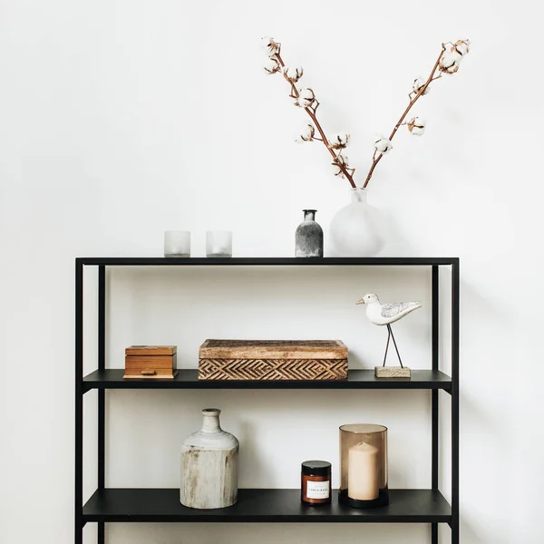 Modern nordic interior design concept. Stylish rack with caskets, cotton branch, vase, candles and bird figurine at white wall.