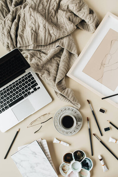 Watercolor painter home office desk workspace on beige background. Coffee cup, laptop, plaid, paint brushes, painting, watercolor. Flat lay, top view art blog, social media, website concept composition.