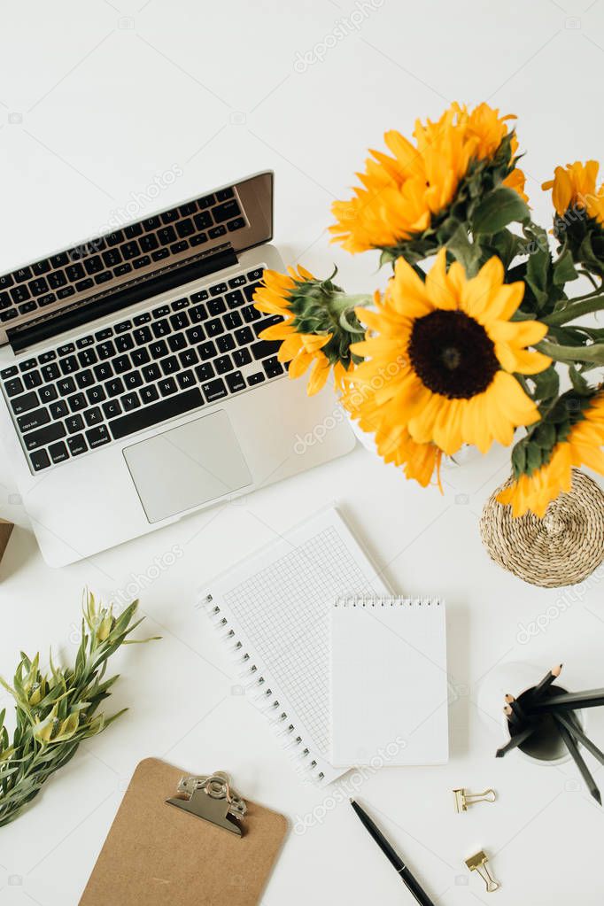 Flatlay of home office desk workspace with laptop, notebook, yellow sunflowers bouquet on white background. Top view freelancer / blogger summer floral work concept.