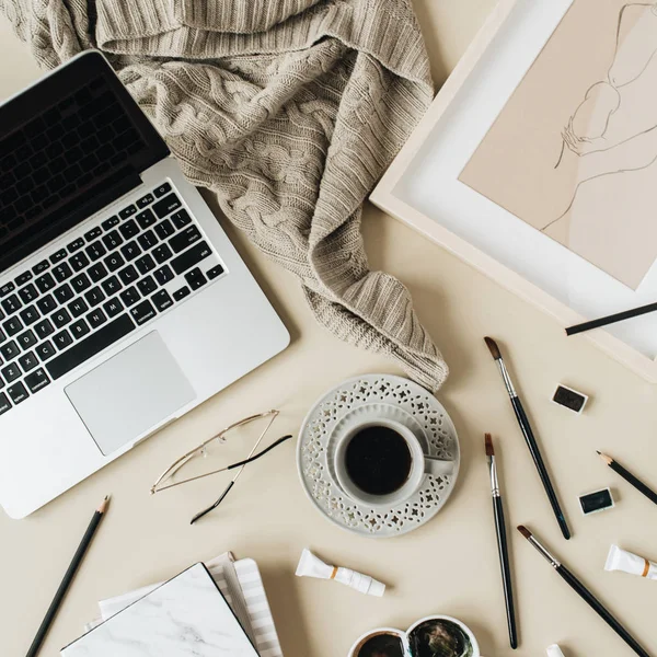 Watercolor painter home office desk workspace on beige background. Coffee cup, laptop, plaid, paint brushes, painting, watercolor. Flat lay, top view art blog, social media, website concept composition.