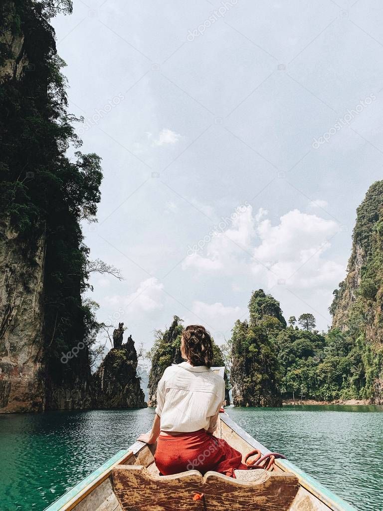 Young woman in red skirt and white blouse sitting on wooden boat watching at exotic and tropical dark green big islands with rocks and turquoise lake at Cheow Lan Lake, Khao Phang, Ban Ta Khun District, Thailand. Travel holiday and adventure concept.